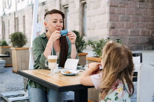 Мать и маленькая дочь едят в уличном кафе. Мать-хипстер с маленькой дочкой обедают вместе в летнем кафе — стоковое фото