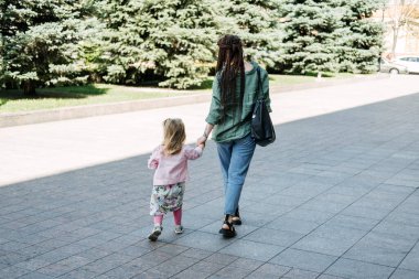 Millennial motherhood, parenthood. Modern hipster mother with dreadlocks and toddler baby daughter walking down the city street
