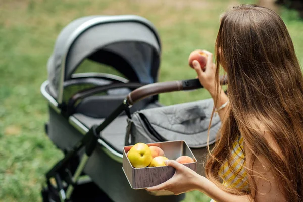 Planes y dieta para madres lactantes. Alergia en un bebé durante la lactancia materna. Joven mamá comiendo frutas mientras camina con el bebé recién nacido en el cochecito. — Foto de Stock