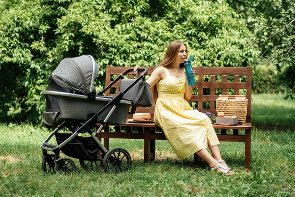 Madre joven con bebé recién nacido en el cochecito bebe agua, sentado en el banco en el parque de verano. Joven madre bebiendo agua después de amamantar al bebé al aire libre — Foto de Stock