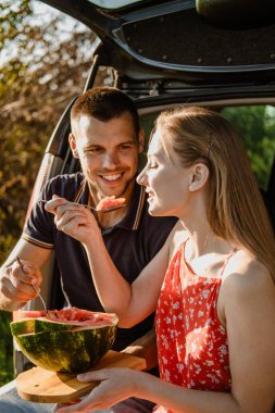 Yerel seyahat, Romantik Piknik Buluşma Fikirleri. Yaz pikniğinde, bagajda karpuzla aşık genç bir çift. Sevgili çift arabanın yanında oturup karpuz yiyor..