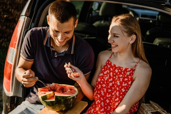 Local travel, Romantic Picnic Date Ideas. Young couple in love on summer picnic with watermelon in car trunk. Loving couple sitting near car and eating watermelon.