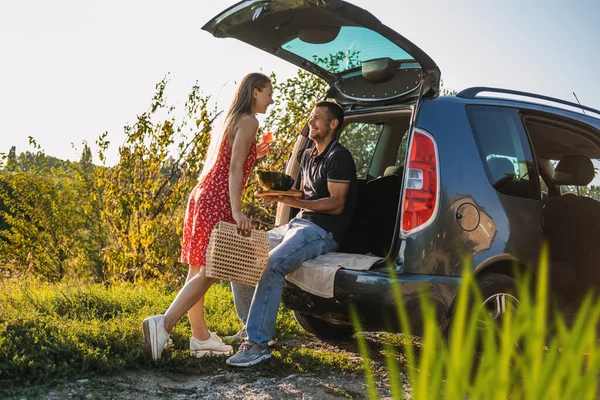 Local travel, Romantic Picnic Date Ideas. Young couple in love on summer picnic with watermelon in car trunk. Loving couple sitting near car and eating watermelon.