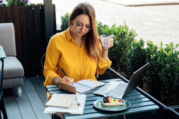 Time management for freelancers. Woman freelancer writes in a notebook and working with laptop in street cafe at summer day.