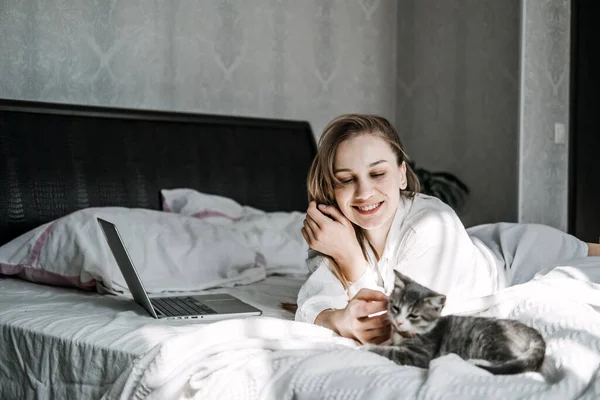 Work from home, remote online work, home office. Woman working on laptop in bed and playing with cute fluffy cat kitten