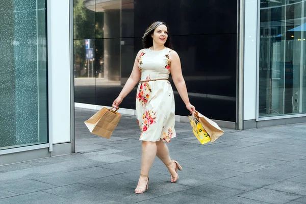 Happy confident smiling plus size curvy young woman with shopping bags and trendy net bag walking on city street near shop mall windows