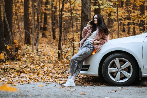 Confident brunette woman standing near car and call phone. Preparing Your Car For Fall autumn season. Driving Tips for Autumn