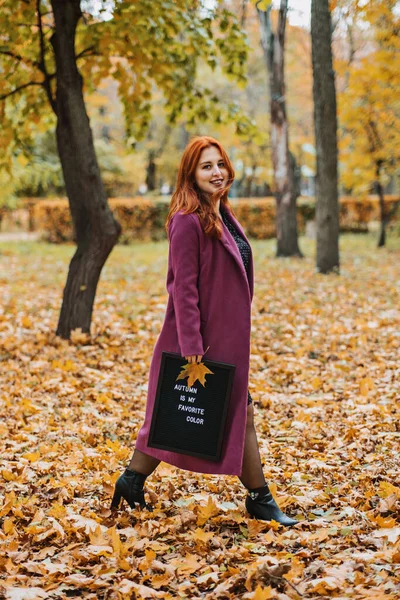 Hello autumn. Red hair girl with Letter message board with text Autumn is my favorite color in hand in fall leaves in park. Autumn motivation quotes