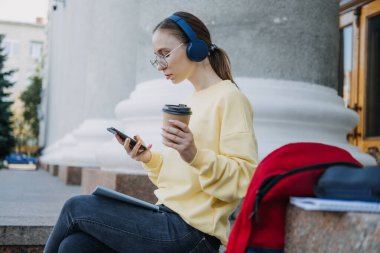 Açık havada cep telefonu uygulaması kullanan kulaklıklı sıradan bir öğrenci portresi. Kulaklık takmış, cep telefonuyla ders çalışan genç bir kız öğrenci. Ses dersi, internet kursu.