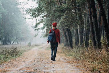 Weekend breaks and getaways in forests. Stay close to nature. Young woman in red hat and sweater with backpack walking in pine forest