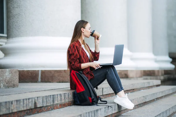 Outdoor portrait of female student with coffee cup and laptop. Girl student has coffee break after lectures, listens audio book in headphones, enjoys record from website, uses mobile phone