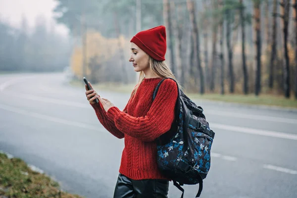 Travel Apps. Young woman traveler with backpack holding smartphone device in hands while walking in pine forest. Happy woman enjoying new adventure and using travel apps or gps navigation in cell