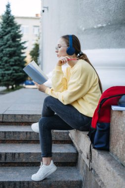 Outdoor Portrait of happy student girl, young woman listening to audio lessons, courses, podcast, music via headphones with book, notebook on the street.