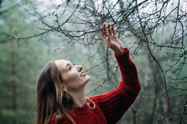 Improving mental wellbeing, Relax and reduce stress. Alone young woman in red sweater touching water drops on tree branches while walking in morning pine forest