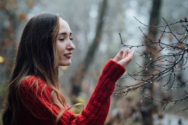 Improving mental wellbeing, Relax and reduce stress. Alone young woman in red sweater touching water drops on tree branches while walking in morning pine forest