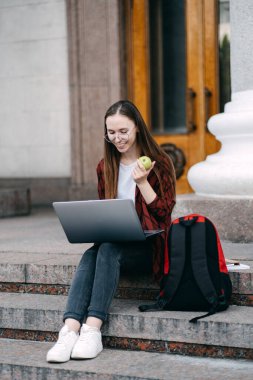 Study snacks to help revise, brain food during exam period. Happy college student girl with apple and coffee cup preparing for exams with laptop and books outdoors