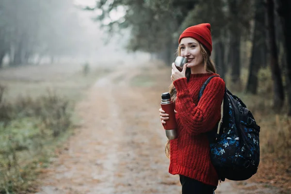 Sustainable tourism, responsible travel. Young woman traveler with backpack holding tremors zero waste cup in pine forest background. Happy woman enjoying new adventure