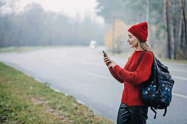 Alone woman with backpack near road on forest background. Hipster girl wanderlust walking on asphalt road in wild lands, female tourist having journey, solo travel
