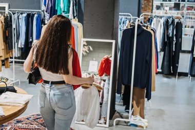Fast fashion, Woman shopping in cloth store. shopping trendy clothes. Young Latina woman standing near hangers with brand wear, spending day in boutique