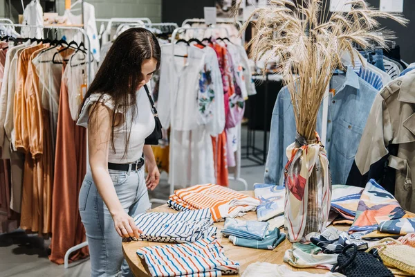 Fast fashion, Woman shopping in cloth store. shopping trendy clothes. Young Latina woman standing near hangers with brand wear, spending day in boutique