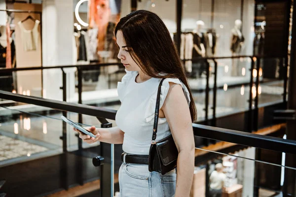 Young Latina woman with shopping bags using her smart phone while standing in shopping center, mall. Woman In Shopping Mall Using Mobile Phone