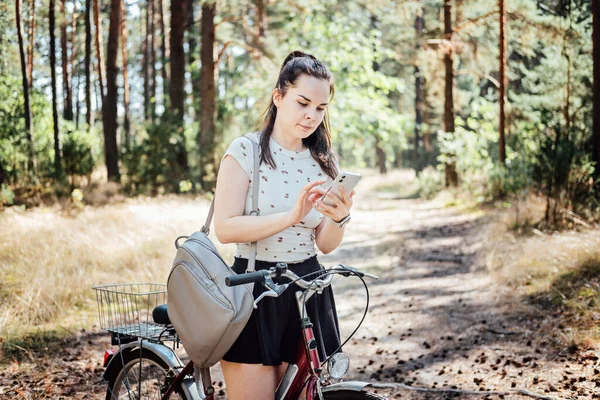 Best cycling apps. Bike Tracker. Young woman with backpack riding bike and looking in cell phone on pine forest background. Girl with bike using a phone texting on smartphone app in forest walk.