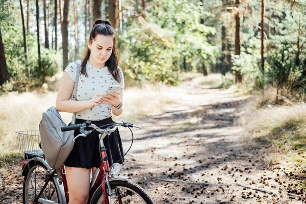 Best cycling apps. Bike Tracker. Young woman with backpack riding bike and looking in cell phone on pine forest background. Girl with bike using a phone texting on smartphone app in forest walk.