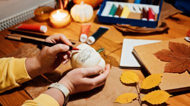 Sonbahar balkabağı zanaatı. Yetişkinler için sonbahar el sanatları. Kadın eli, beyaz balkabağına sonbahar dekoruna karşı evdeki masada desenler çizer..