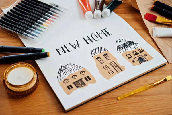 Dreaming about new house. Drawing of several houses text new home on table with paints, brushes and stationery, supplies for drawing and craft.