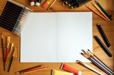 Yaratıcı sanatçı, ahşap masadaki iş yerinde maket yapıyor. Düz yatıyordu, üstü boş eskiz defteriyle telif alanı, kalemler, boya fırçaları ve boyalarla doluydu. Sanat, atölye, resim, çizim, zanaat, yaratıcılık