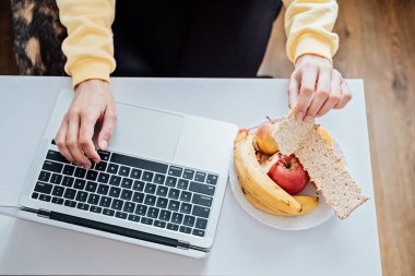 Evde çalışırken sağlıklı beslenen serbest çalışan genç bir kadın. Ev bilgisayarında çalışırken sağlıklı tahıllı atıştırmalık ve meyve yiyen bir kadın..