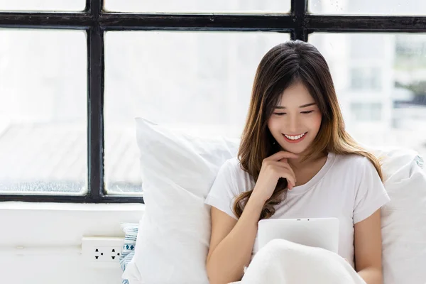 笑的年轻亚洲女人在卧室里用平板电脑在平板电脑里打扫卫生快乐漂亮的女孩总是用平板电脑在网上购物和看电影 — 图库照片
