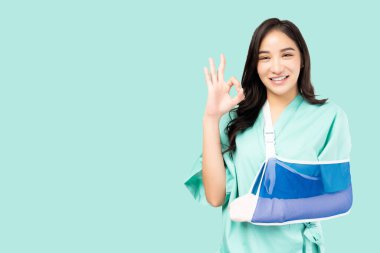 Beautiful young asian woman wearing patient gown and soft splint and wrapped in bandages on arm showing okay sign on green background Patient Insurance Health care and Personal accident concept