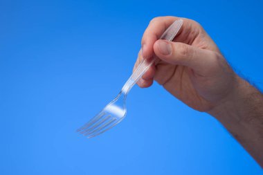 Tek kullanımlık şeffaf plastik çatal insan eliyle tutulur. Stüdyo çekimini kapat, mavi arkaplanda izole edilmiş..