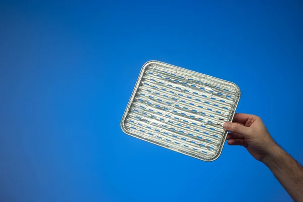 Empty single use metal aluminum foil cooking tray held by Caucasian male hand. Close up studio shot, isolated on blue background.