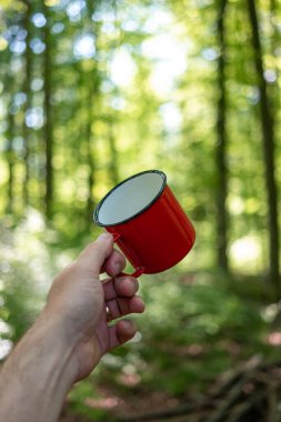 Kızıl metal kamp ya da piknik kupası ya da beyaz bir erkeğin elinde tuttuğu bardak. Gündüz yeşili orman düzeni, sığ alan derinliği.