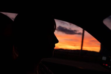 Arabasını kullanan genç beyaz bir kadının siluet portresi. Dramatik bir gün batımına karşı gökyüzü ve bulutlar, tanınmayan bir yüz.