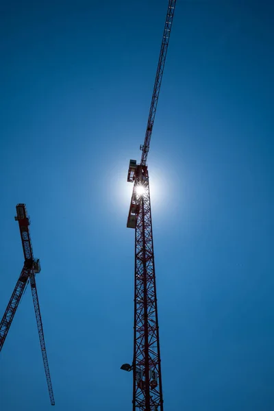 A pair of construction cranes set against the noon sun. Blue sky, no clouds, low angle, no people, silhouette.