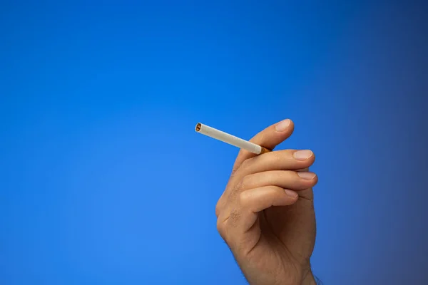 An unlit cigarette held in hand by Caucasian male hand. Close up studio shot, isolated on blue background.