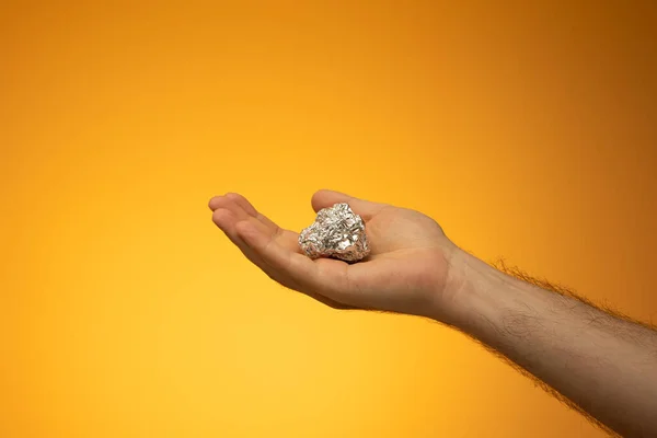 Kitchen tin foil crumpled into a ball held in hand by Caucasian male hand. Close up studio shot, isolated on orange background.