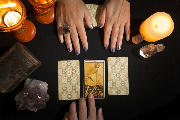 Bir kadının elinde siyah kartlardan oluşan bir masada üç tarot kartı gösterirken, diğer elinde seçilmiş kişi olarak ölüm kartı görülüyor. Tarot kartlı kehanet oturumu. Yukarıdan görüntüle.