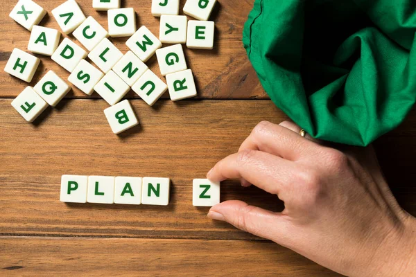 A hand placing a piece with the letter Z next to the word PLAN. An empty green bag and other pieces with letters appear displaced. Concept of planning for the future, endurance and constancy.