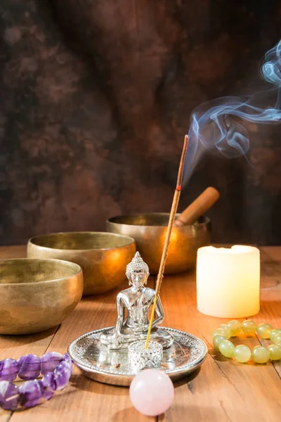 Still life with singing bowls, minerals, a candle, incense and a Buddha figure on wooden boards and a dark background. Small candle-lit altar for meditation and music therapy.