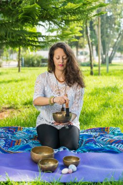 Yoga minderlerinin üzerinde diz çökmüş bir kadın park alanında meditasyon ve müzik terapisi sırasında Tibet şarkı kasesi çalıyor.