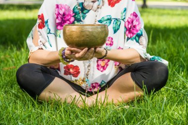 Detail of a woman's hands holding a Tibetan singing bowl, sitting on the grass during a meditation and music therapy session in a park.