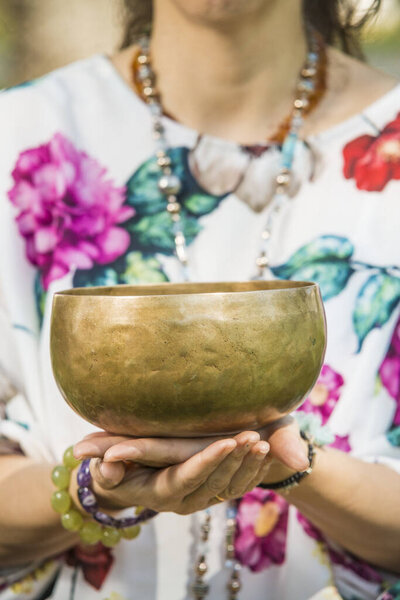 Detail of a woman's hands holding a Tibetan singing bowl during a meditation and music therapy session in a park area.