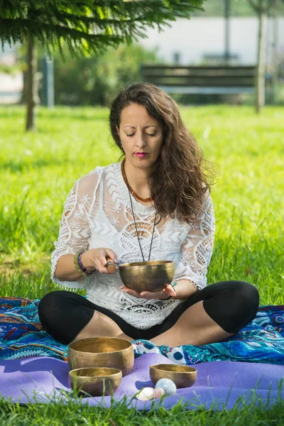 A woman, seated on yoga mats, plays a Tibetan singing bowl during a meditation and music therapy session in a park area.