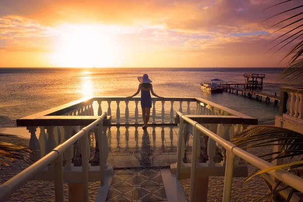 Une femme dans un chapeau regardant le coucher de soleil romantique des Caraïbes tout Image En Vente