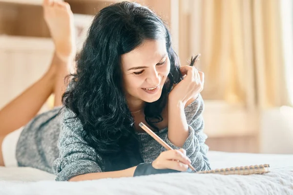 Mujer atractiva feliz sonriendo y dibujando con lápiz en el cuaderno mientras está acostado en el sofá —  Fotos de Stock