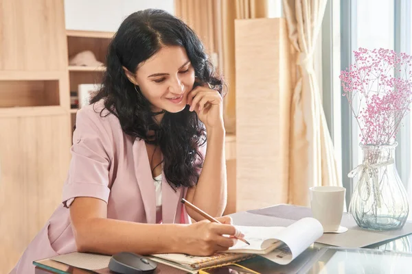 Mujer atractiva joven sonriendo y dibujando con lápiz en el cuaderno mientras está sentado en la mesa Imágenes De Stock Sin Royalties Gratis